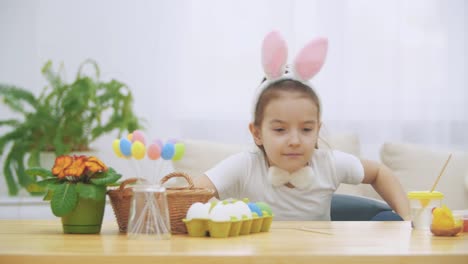 Little-nice-girl-is-climbing-into-a-soft-chair,-and-is-playing-with-bunny.-She-sits-down-at-the-table-with-Easter-decorations-and-is-painting-a-Bunny.