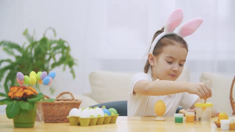 Little-playful-girl,-wearing-bunny-ears-on-her-head-is-choosing-an-a-red-colour-to-paint-an-egg.