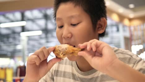 Happy-Asian-preteen-boy-eating-fried-chicken-leg-in-fast-food-restaurant.