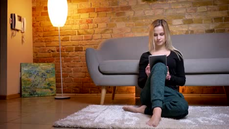 Closeup-portrait-of-young-pretty-blonde-haired-female-using-the-tablet-sitting-on-the-floor-carpet-indoors-in-a-cozy-apartment