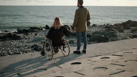 Woman-is-sitting-in-wheelchair-on-sea-coast-and-holding-hand-of-her-husband