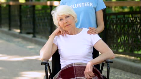 Volunteer-with-upset-senior-woman-in-wheelchair-looking-at-camera,-hospital-park