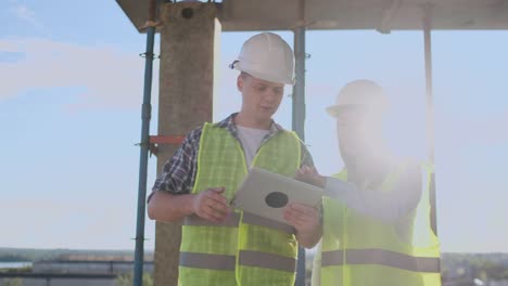 Trabajadorde-la-construcción-hombre-y-arquitecto-mujer-en-un-casco,-discutir-el-plan-de-construcción-de-la-casa,-hablarse-unos-a-otros-sobre-el-diseño,-sosteniendo-una-tableta,-mirar-los-dibujos,-fondo-de-los-rayos-del-sol