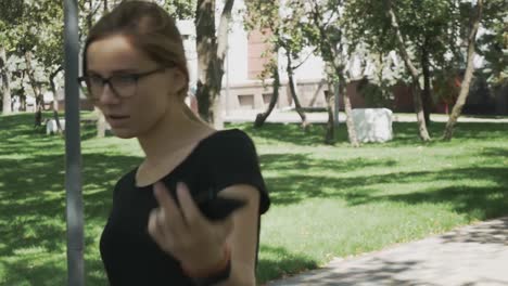 Mujer-caucásica-bastante-joven-en-anteojos-caminando-hacia-la-cámara,-revisando-el-teléfono-y-celebrando-buenas-noticias-en-un-parque