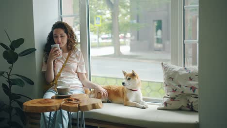 Chica-bonita-usando-teléfono-inteligente-acariciando-lindo-perro-sentado-en-el-alféizar-de-la-ventana-en-el-café