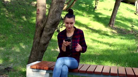 Young-beautiful-girl-in-a-plaid-shirt-is-sitting-on-a-park-bench,-smiling-and-writing-a-message-in-her-smartphone.