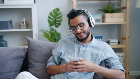 Guapo-árabe-chico-usando-teléfono-inteligente-escuchando-música-en-auriculares-en-casa