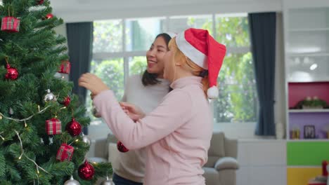 Asiatische-Freundinnen-schmücken-Weihnachtsbaum-auf-Demonstaus.-Weibliche-teen-glücklich-lächeln-feiern-Weihnachts-Winterurlaub-zusammen-im-Wohnzimmer-zu-Hause.-Zeitlupenaufnahme.