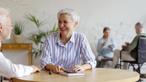 Tracking-shot-of-elderly-couple-sitting-at-table-and-having-fun-talking-joyfully,-senior-woman-and-disabled-senior-man-in-wheelchair-playing-cards-in-background-in-assisted-living-home