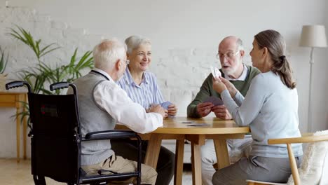 Tracking-shot-of-group-of-four-retired-elderly-people,-two-men-and-two-women,-sitting-at-table-and-playing-cards-together-in-common-room-of-assisted-living-home