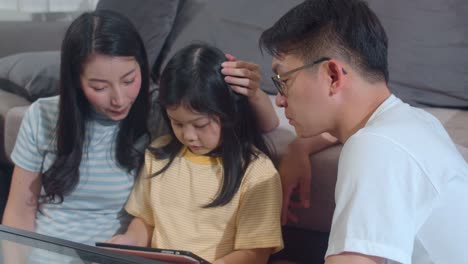 Joven-familia-asiática-e-hija-feliz-usando-la-tableta-en-casa.-Madre-japonesa,-padre-relajarse-con-la-niña-viendo-películas-tumbada-en-el-sofá-de-la-sala-de-estar.-Padre-divertido-y-niño-encantador-se-están-divirtiendo.