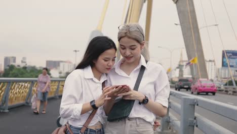 Asian-lesbian-couples-using-smartphones-browsing-the-website-while-standing-on-the-bridge.-Low-angle-shot.