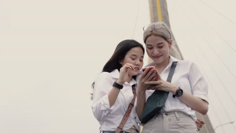 Asian-lesbian-couples-using-smartphones-browsing-the-website-while-standing-on-the-bridge.-Low-angle-shot.