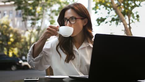 Weiblich-in-Brille,-weißes-Hemd.-Sie-sitzt-mit-Laptop-und-Smartphone-am-Tisch-in-einem-Straßencafé,-trinkt-Kaffee-und-arbeitet-online.-Nahaufnahme