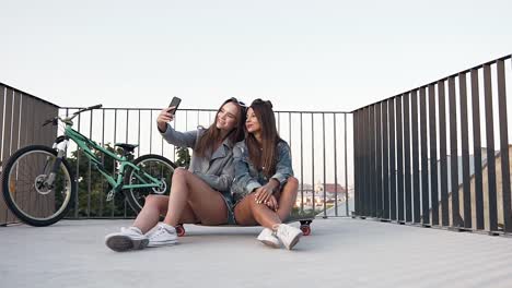 Chicas-sexy-bastante-jovial-sentadas-en-el-monopatín-y-haciendo-selfie-con-caras-divertidas-en-la-terraza-de-la-azotea