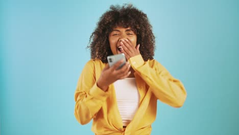 Afro-american-lady-looking-at-smartphone,-saying-yes,-raising-fists-up-being-surprised-and-overjoyed,-smiling-posing-on-blue-background