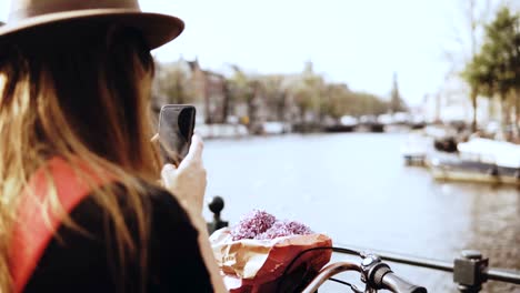 Tourist-lady-with-bicycle-takes-photos-on-bridge.-Female-with-long-hair-and-flowers-photographs-beautiful-river-scenery