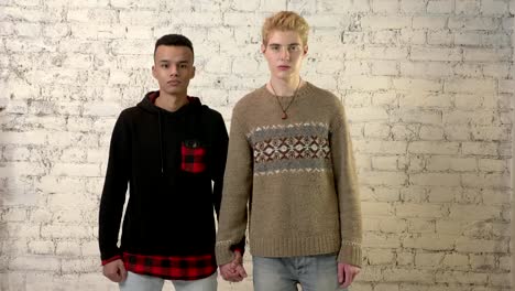 Sad-International-gay-couple-holding-hands,-white-brick-wall-on-background.-LGBT-love,-young-couple,-lover,-homosexuality-concept.-60-fps