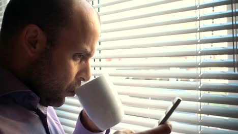 Tired-Businessman-standing-with-smartphone-by-window-in-the-office.-Close-up,-serious-man-sitting-by-out--window-and-uses-a-smartphone.-Thoughtful,--problem,-social-network.-Boss-drinks-coffee,-tea.