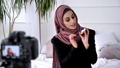 Hermosa-India-joven-en-blogger-hijab-hablando-en-la-cámara,-sonriendo,-mostrando-una-nueva-compra,-cosméticos,-aquagrum,-confort-en-el-hogar-en-el-fondo.-60-fps