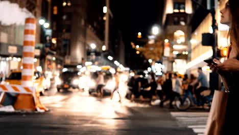 Young-beautiful-woman-crossing-the-traffic-road-in-downtown-of-New-York,-America-in-the-evening-and-using-smartphone