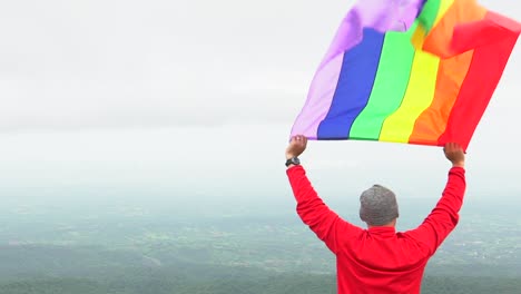 Mann-erhöhen-Regenbogen-Farbe-LGBTI-Fahnenschwingen-in-harten-Wind-auf-Mountain-Top-Sicht