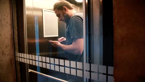 Happy-casual-Caucasian-male-freelancer-riding-transparent-glass-elevator,-door-opens-and-he-walks-out-using-smartphone