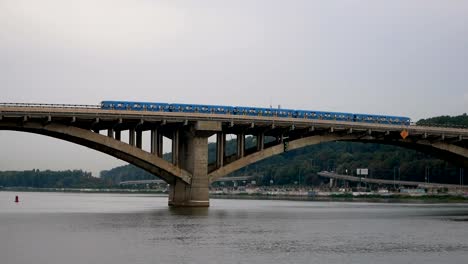 Zug-fährt-über-die-Brücke-gegen-den-Himmel-und-den-Wald.-City-u-Bahn-links-außen.-Öffentlichen-Verkehrsmitteln-unterwegs.-Autos-fahren-über-die-Brücke-über-den-Fluss.