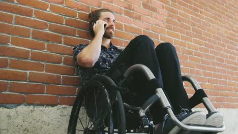 Joven-discapacitado-realizar-llamada-telefónica-en-silla-de-ruedas-inclinada-contra-la-pared-de-ladrillo