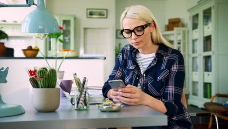 Attraktive-blonde-Geschäftsfrau-SMS-auf-Smartphone-im-Home-Office