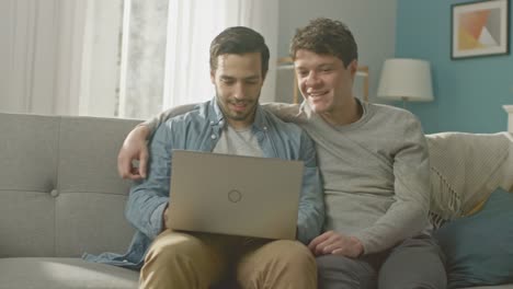 Adorable-Pareja-Gay-hombre-pasar-tiempo-en-casa.-Se-sientan-en-un-sofá-y-usar-la-Laptop.-Navegan-en-línea.-Mano-de-socio-es-alrededor-de-su-amante.-Habitación-tiene-un-Interior-moderno.