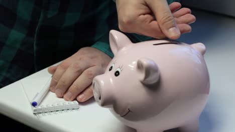 On-the-table-there-is-a-piggy-bank-in-the-form-of-a-pink-pig.-A-man-throws-coins-at-her.-Makes-notes-in-a-notebook.