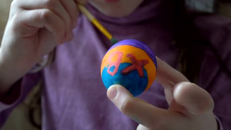 A-little-girl-holds-an-Easter-egg-in-her-hand-and-paints-on-it-a-brush-and-colored-paints,-sitting-at-home-at-the-table.-Close-up.-4K.