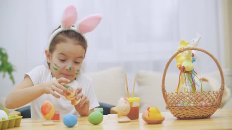 Nice,-cute-girl-is-having-fun-painting-an-Easter-egg.-Adorable-girl-is-colorizing-Ester-egg.-Girl-with-beauty-spots-at-her-face-and-is-smiling-gently-with-cat's-whisker,-sitting-at-the-wooden-table-with-Easter-decorations.-Gently-smiling-little-girl-shows