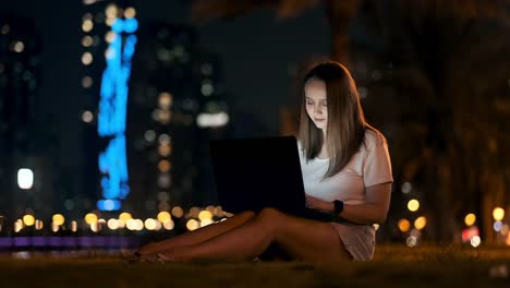 Jovencita-noche-de-verano-sentada-en-la-hierba-con-Laptop-en-la-ciudad