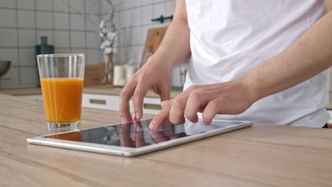 Hombre-atractivo-en-casa-usando-la-tableta-en-la-cocina-enviando-mensaje-en-las-redes-sociales-sonriendo-disfrutando-del-estilo-de-vida-moderno-que-llevaba-camisa-blanca