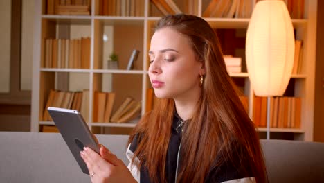 Portrait-of-young-teenage-girl-searching-in-tablet-sitting-on-sofa-on-bookshelves-background-watches-into-camera-at-home.