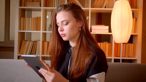 Portrait-of-young-teenage-girl-watches-into-tablet-sitting-on-sofa-on-bookshelves-background-at-home.