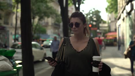 Atractiva-mujer-caucásica-con-gafas-de-sol,-pecas,-piercings-y-pelo-rojo-desplazando-las-redes-sociales-en-su-teléfono-inteligente-caminando-por-la-calle,-durante-el-verano-soleado-en-París.-4K-UHD.-Moda.