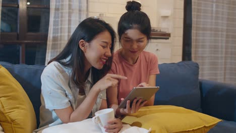 Lesbian-lgbt-women-couple-using-tablet-at-home,-Asian-female-happy-using-technology-check-social-media-in-internet-together-while-lying-sofa-in-living-room-in-night.-Lover-celebrate-holiday-concept.