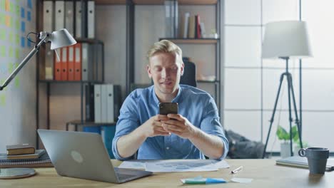 Creative-Entrepreneur-Sitting-at-His-Desk-Works-on-a-Laptop-in-the-Stylish-Office,-Uses-Smartphone-for-Social-Media-Apps,-Emailing-Business-Associates,-Reading-News,-Browsing-through-Internet