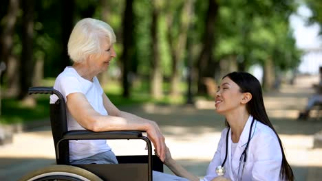 Smiling-nurse-talking-with-disabled-elderly-woman-in-park,-rehabilitation-center