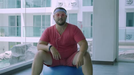 Lustige-fette-Männchen-in-rosa-Brille-und-in-einem-rosa-T-Shirt-ist-auf-einem-fit-Ball-in-der-Turnhalle-darstellung-ein-Mädchen-engagiert.-Ein-wunderbarer-Mann-hört-Musik-und-tanzt-auf-dem-Ball-in-der-Turnhalle