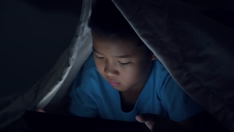 Little-boy-using-tablet-under-blanket-at-night-in-bed.