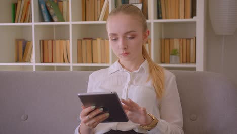 Retrato-de-primer-plano-de-joven-bonita-estudiante-rubia-caucásica-usando-la-tableta-mirando-la-cámara-sentado-en-el-sofá-interior-en-el-apartamento