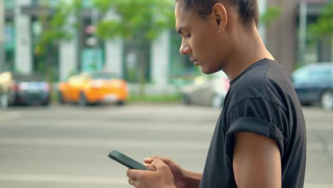 perfil-hombre-afroamericano-caminando-sosteniendo-teléfono-inteligente-a-lo-largo-de-la-calle-en-la-ciudad