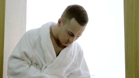 Man-dressed-in-white-bathrobe-sitting-on-windowsill,-using-tablet-computer.-4K