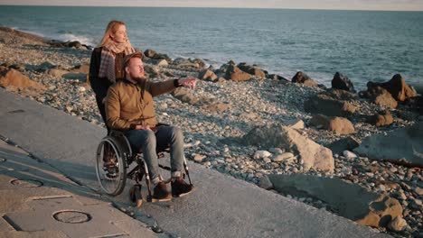 Young-woman-is-nursing-sick-disabled-man,-rolling-stroller-on-sea-shore