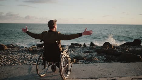 Male-disabled-is-watching-sea-horizon-and-lifting-hand-up,-back-view