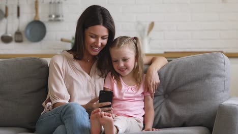 Happy-mom-and-child-daughter-enjoying-using-smartphone-on-sofa